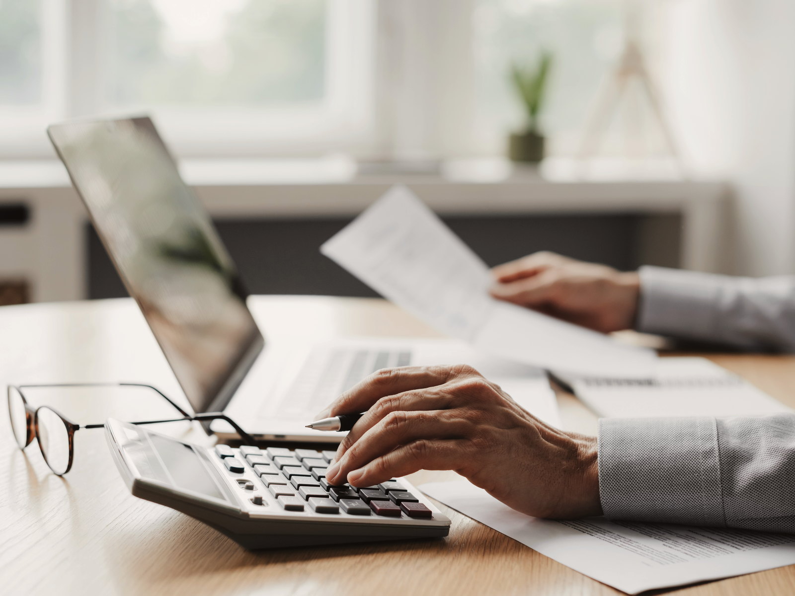 Hands typing on a laptop beside a calculator and printed statements on a bright desk