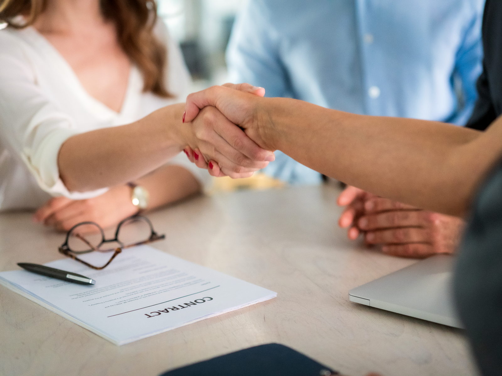 Two people shaking hands across a desk over a contract, glasses and a pen beside it